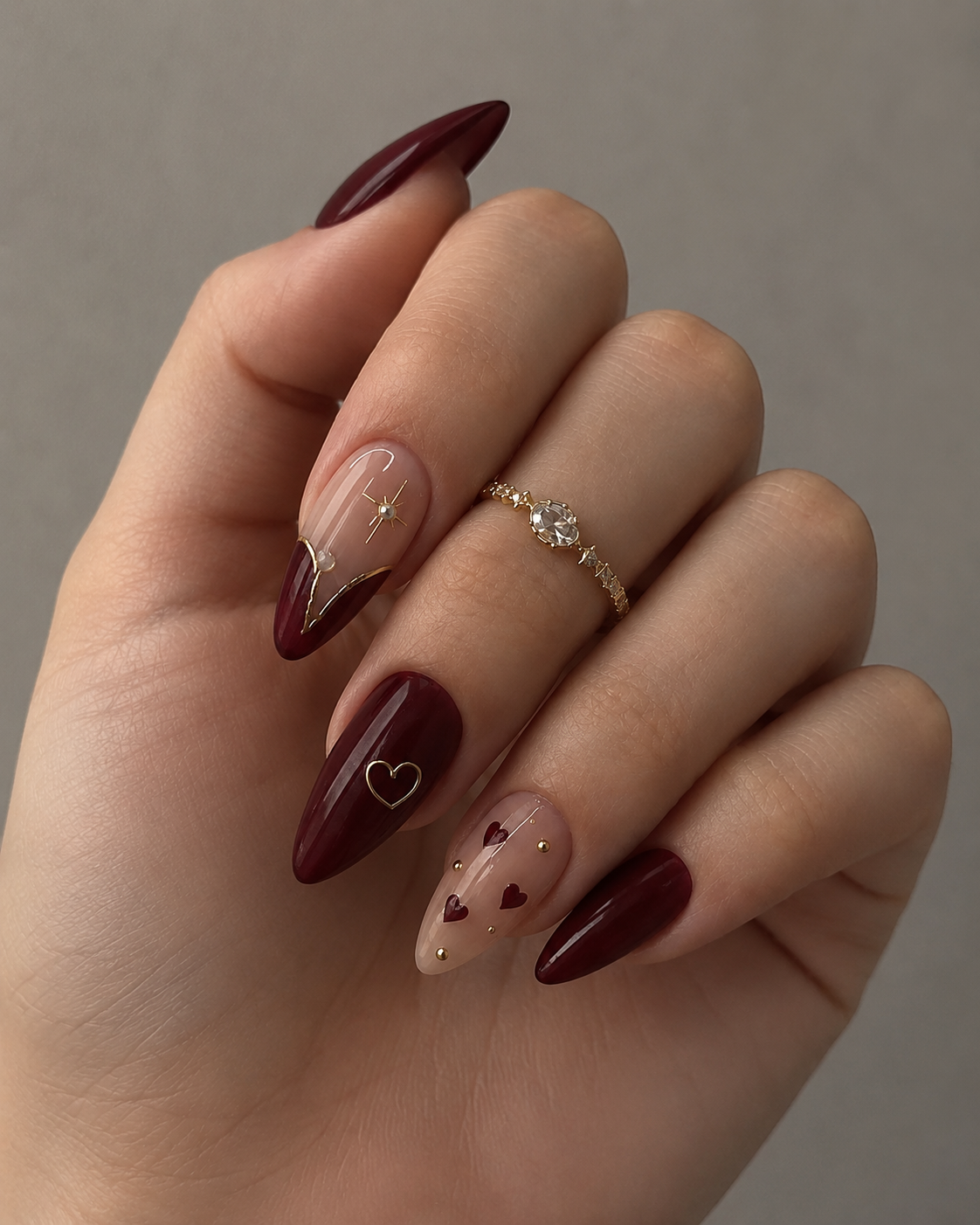Deep burgundy almond nails showcasing a celestial and romantic theme. The index finger features a gold-rimmed reverse French tip with a starburst, while other nails are accented with gold hearts, mini red hearts, and delicate gold studs.