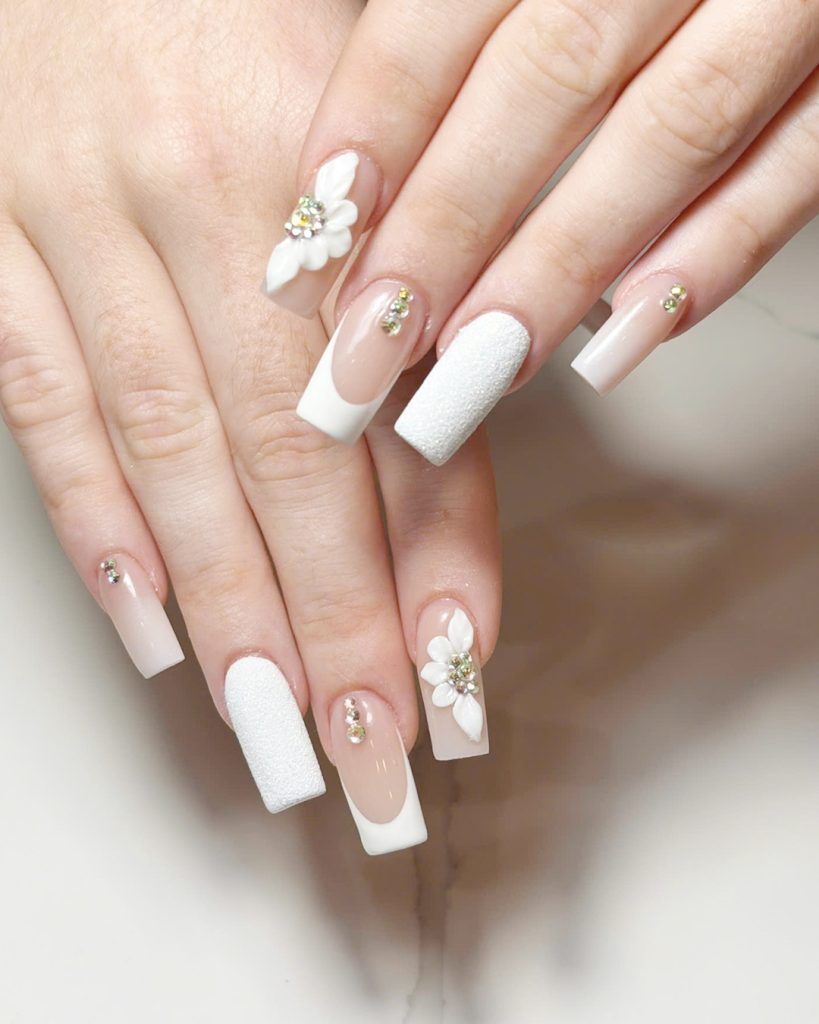 Builder Gel Nails vs. Acrylic: What Changes When You Switch 4 Long square nude nails with white French tips, sugar-textured white accents, 3D white flowers and rhinestones