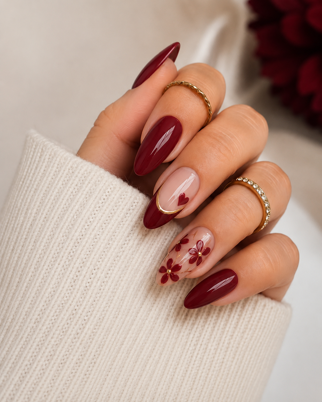 Glossy deep burgundy almond nails featuring a cut-out heart design framed in gold foil, accompanied by delicate red floral painting on a sheer nude accent nail.