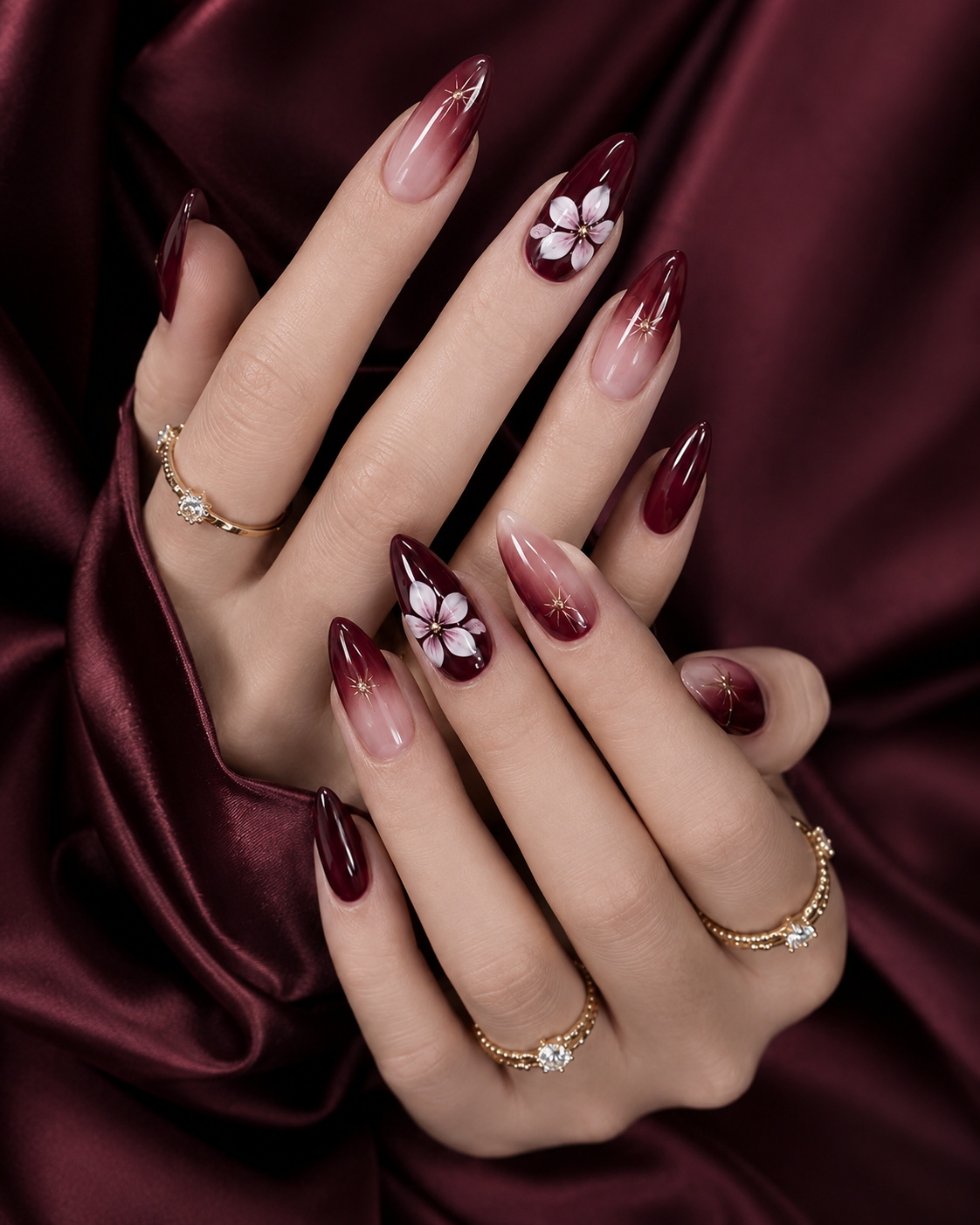 Dark burgundy ombre almond nails blending from a deep vampy red tip to a soft nude base, decorated with white 3D floral accents and delicate gold starburst details.