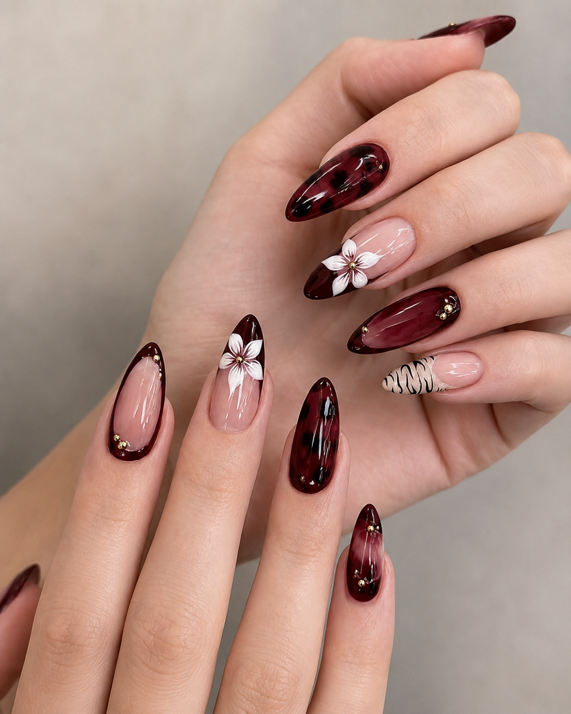 Vampy red tortoiseshell-effect almond nails mixed with fine line animal print accents, French tip outlines, gold studs, and white floral art.