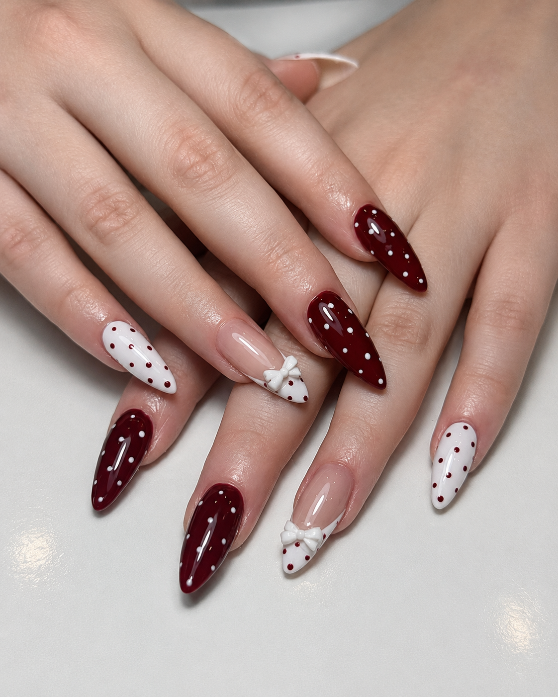 Almond nails with a bold white-and-burgundy polka dot pattern, featuring white French tips decorated with 3D white bow charms on a natural nude base.