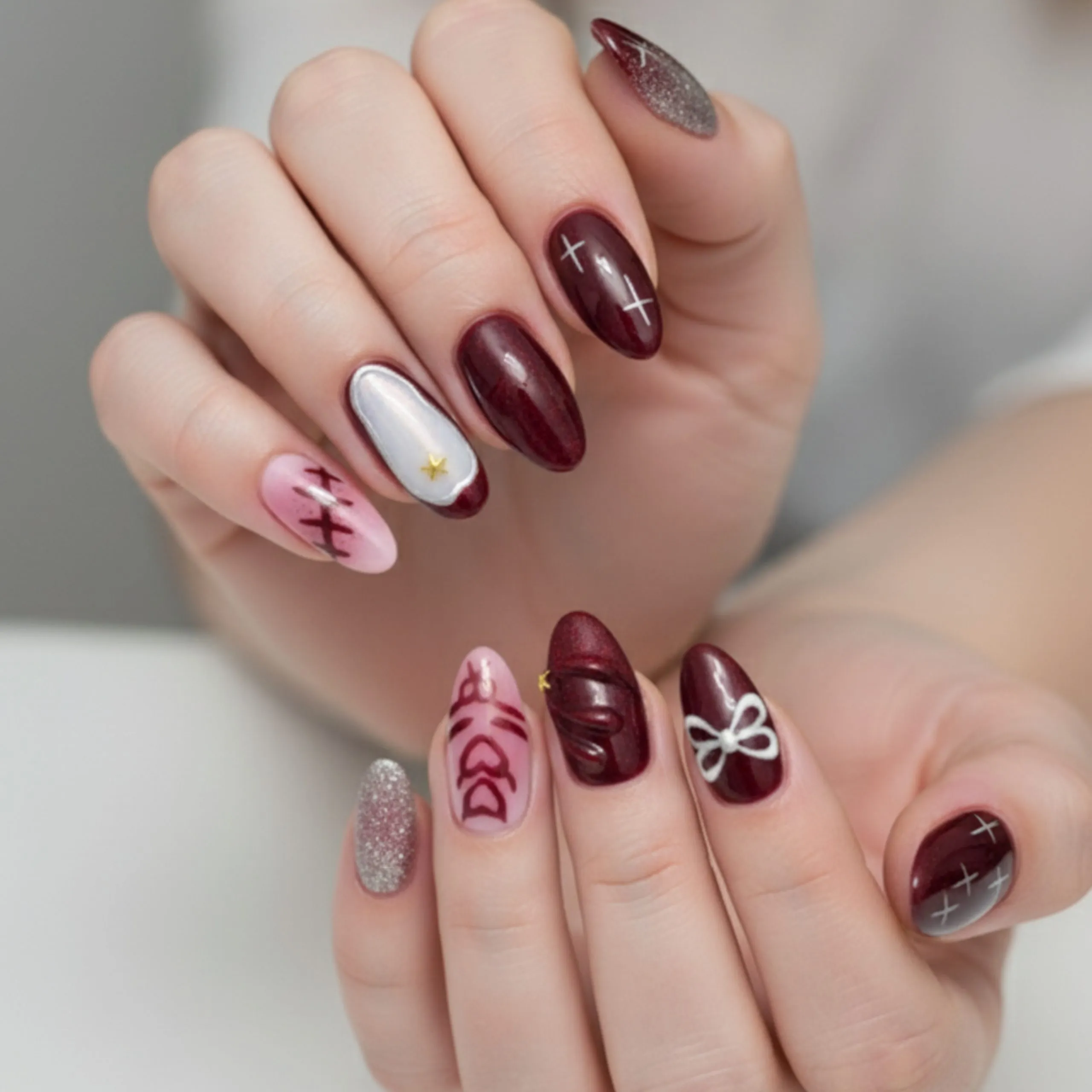 7 Homecoming Nails Styles to Match Every Personality 2 Bold homecoming nails with deep burgundy and glitter accents, featuring 3D ribbon art, abstract face design, and silver cross detailing on oval-shaped nails.