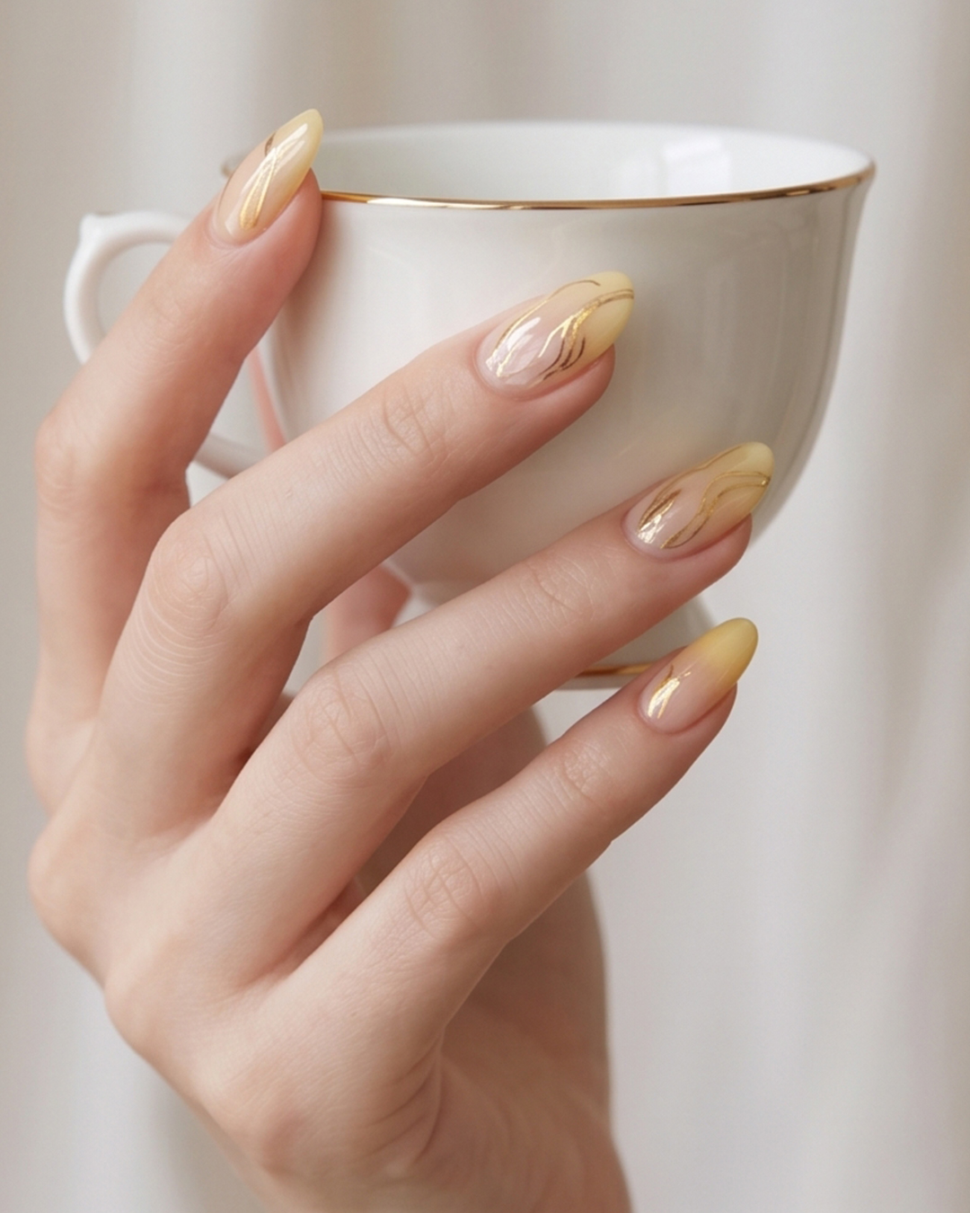 Butter yellow nails with gold accents and metallic swirl nail art on almond-shaped nails.