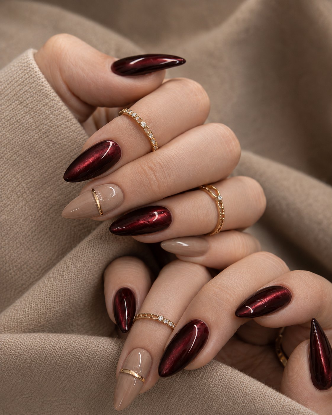 Long almond burgundy cat eye nails alternating with sheer nude nails and stacked gold rings