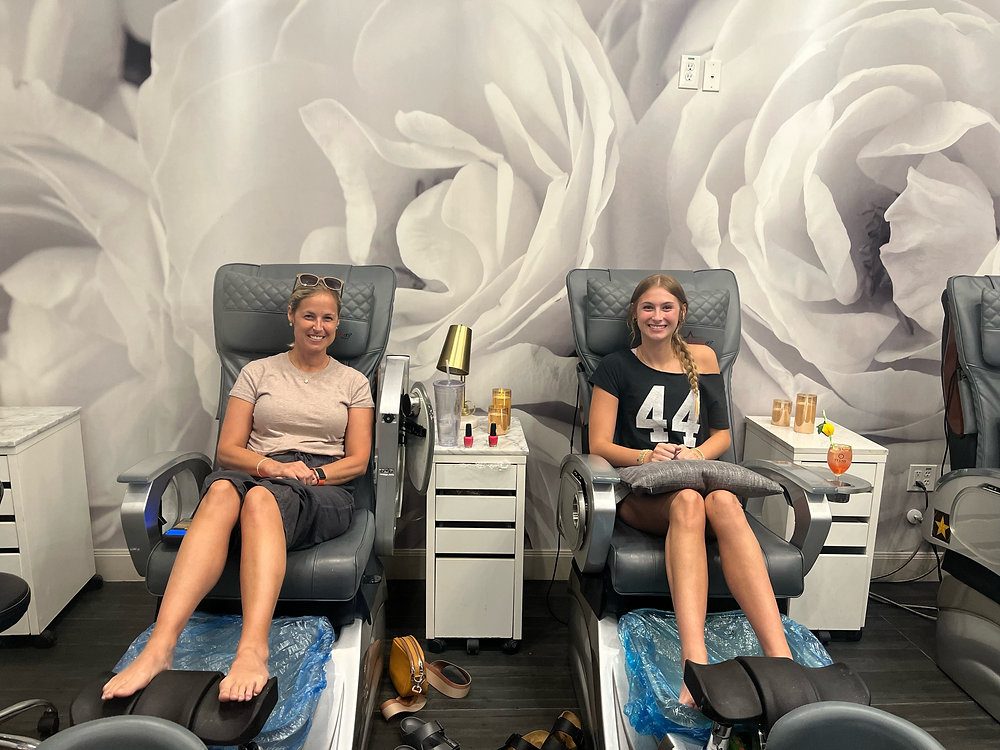 Two smiling female clients enjoying a relaxing day at Plush Nail Bar, seated in modern grey pedicure massage chairs. They are receiving specialized foot spa treatments in a clean, luxury salon environment with white rose mural wallpaper.
