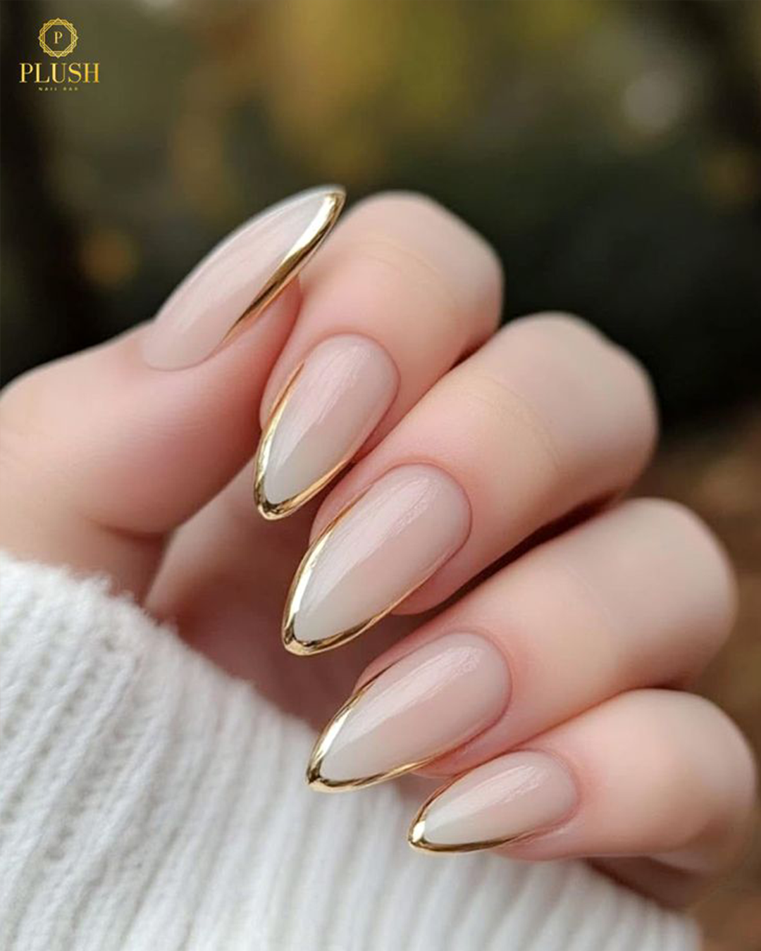 Close-up of nude almond nails with shiny metallic gold French tips at Plush Nail Bar.