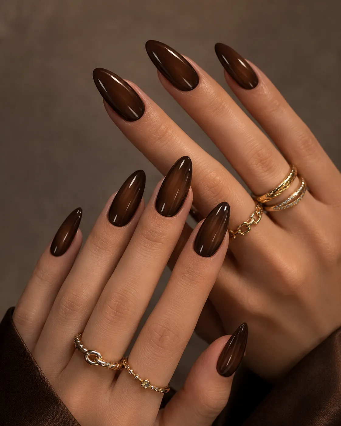 8 Ways to Wear Brown Nails This Season in Houston 4 Glossy brown almond-shaped nails on hands with layered gold rings