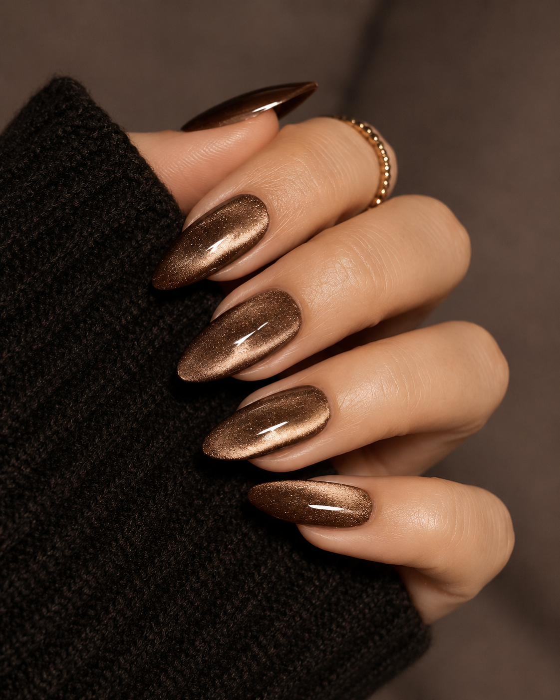 Hand with long shimmery brown almond nails and a gold ring, wearing black sweater