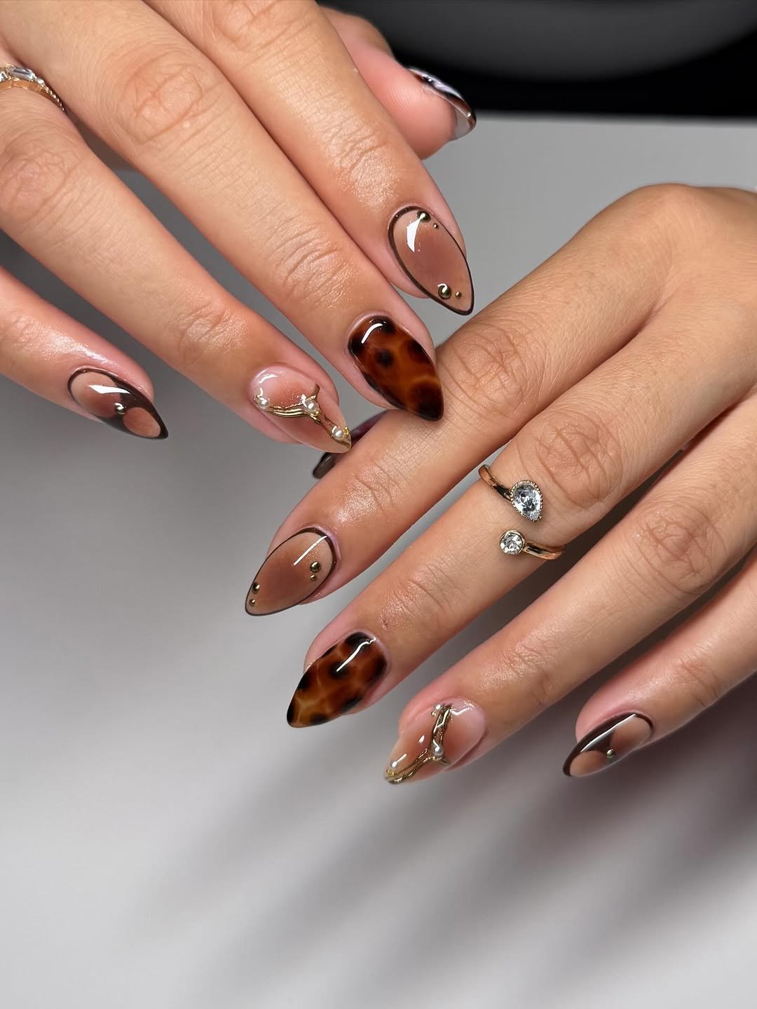 Sheer brown jelly almond nails outlined in dark brown with tortoiseshell accents, gold studs and a gold charm
